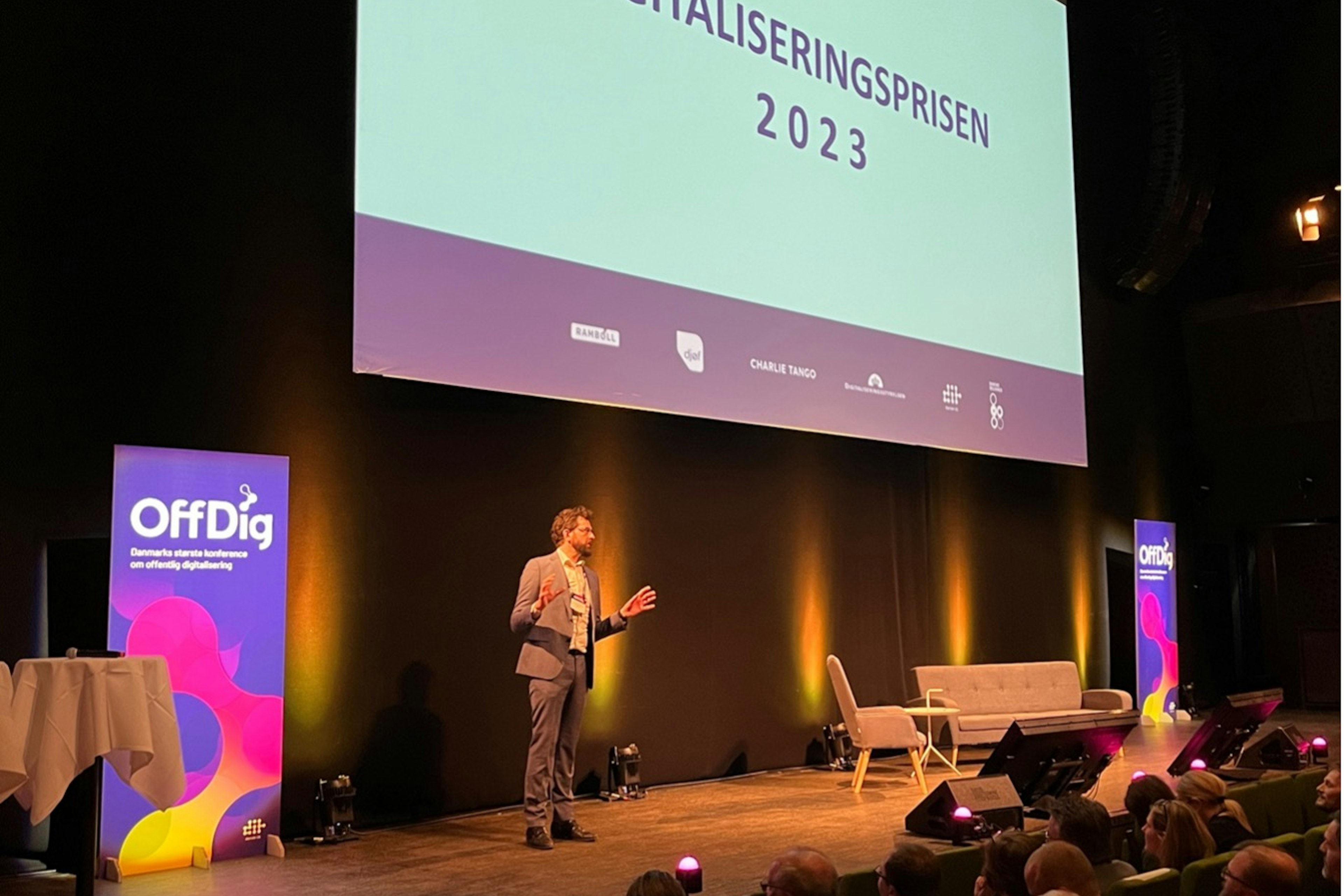 Mand stående på scenen til Digitaliseringsprisen 2023. Publikum sidder foran. Sofa og stol stående på scenen og skilte med 'Offdig' skrevet på.